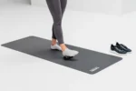 Pies con calzado deportivo sobre un rodillo Blackroll Mini claro, en el suelo junto a un mat