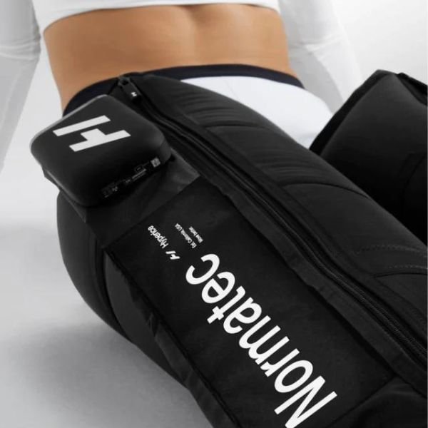 Vista cercana de una bota Normatec Elite puesta en la cadera de una persona, mostrando la cremallera, el texto Normatec y el logo Hyperice en la unidad de control