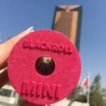 Mano sosteniendo el rodillo Blackroll Mini rosado al aire libre, con un edificio de fondo