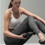 Mujer sentada en el suelo usando una pistola de masaje negra sobre su pierna, vistiendo top deportivo blanco y calzas grises