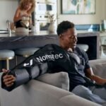 Hombre sentado en un sillón usando mangas de compresión Normatec en ambos brazos, ambiente doméstico de fondo