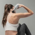Mujer usando una pistola de masaje sobre su propio hombro, vista de espaldas, con coleta y top deportivo blanco