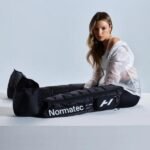 Mujer sentada junto a ambas botas Normatec Elite abiertas y extendidas sobre una superficie, mostrando el interior y el logo Hyperice