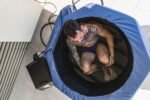 Vista aérea de un hombre sentado dentro de la Bañera Nubis Ice Bath de tela azul, sumergido hasta la cintura, con el chiller-bomba negro junto a la bañera sobre piso de cerámica