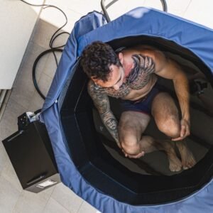 Vista aérea de un hombre sentado dentro de la Bañera Nubis Ice Bath de tela azul, sumergido hasta la cintura, con el chiller-bomba negro junto a la bañera sobre piso de cerámica