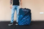 Hombre de pie junto al bolso de transporte azul con estampado geométrico y logo Nubis de la Bañera Ice Bath, apoyado sobre una colchoneta negra