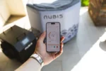 Mano sosteniendo un teléfono con la app Nubis mostrando el control de temperatura, con la Bañera Ice Bath cubierta y el chiller negro de fondo en exterior