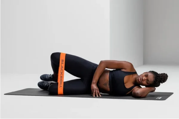 Mujer haciendo ejercicio de clamshell con Blackroll Loop Band naranja sobre las rodillas