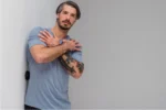 Hombre con tatuajes en el antebrazo, apoyado contra una pared, sosteniendo el Trigger negro sobre el hombro para trabajar un punto gatillo