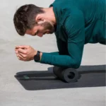 Hombre en plancha con el antebrazo apoyado sobre un rodillo, vistiendo polerón verde