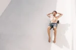 Hombre sin polera recostado sobre una superficie clara, con las manos detrás de la cabeza, estirando el torso al sol