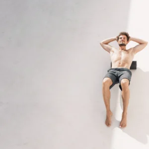 Hombre sin polera recostado sobre una superficie clara, con las manos detrás de la cabeza, estirando el torso al sol