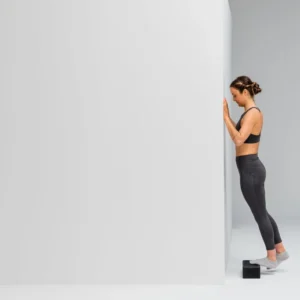 Mujer haciendo elevación de talón apoyada contra la pared con el Blackroll Block bajo el pie